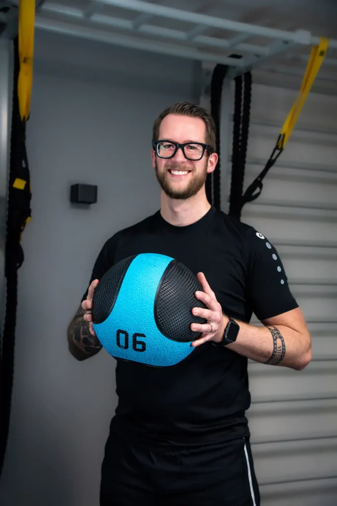 Thomas Adel, Geschäftsführer und Personal Trainer bei SOMT!X, hält lächelnd einen blauen Medizinball.